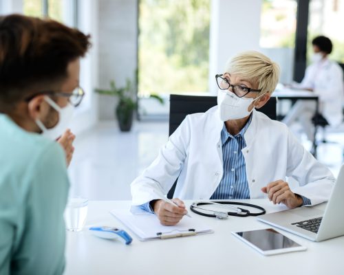 Mature female doctor wearing protective face mask while having appointment with a patient at medical clinic during coronavirus epidemic.